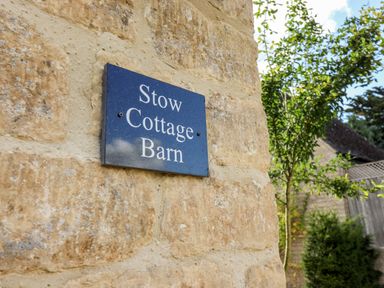 A sign reading Stow Cottage Barn on a stone wall with trees and a fence in the background at Stow Cottage Barn in Churchill Oxfordshire