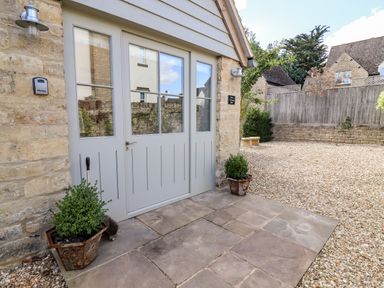 A stone house entrance with a grey door flanked by two potted plants on a stone patio at Stow Cottage Barn in Churchill Oxfordshire