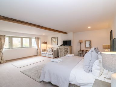 A bedroom with a bed towels on it a striped sofa a television on a patterned cabinet windows with curtains and wooden beam ceiling at Stow Cottage Barn in Churchill Oxfordshire