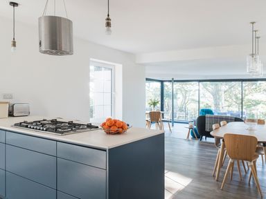A kitchen with a gas stove and oranges on the counter next to a dining area with wooden chairs and large windows showing trees at Landmark in Salcombe