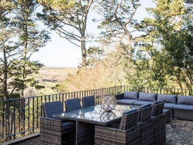 An outdoor patio with wicker chairs around a glass table and a sectional sofa surrounded by trees at Landmark in Salcombe