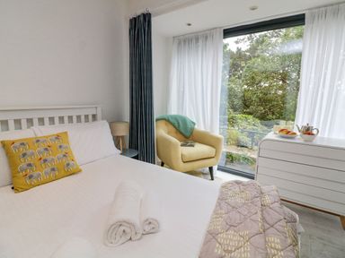 A bedroom with a bed with rolled towels and an elephant pillow a yellow armchair near a large window and a white dresser with breakfast items at Landmark in Salcombe