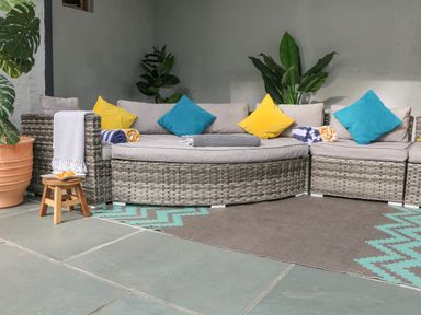An outdoor seating area with a wicker sectional sofa with cushions and colorful pillows a small wooden stool with a dish and a blanket draped over the sofa at Landmark Salcombe