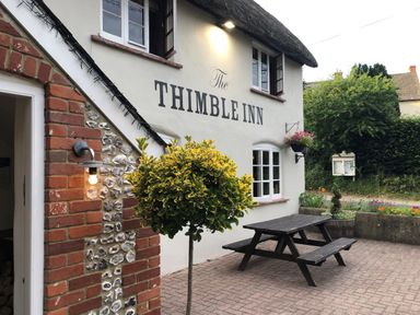 Exterior of The Thimble Inn with a black picnic table a round bush and hanging flower basket