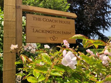 A wooden sign with the text The Coach House at Lackington Farm surrounded by green plants and pink flowers