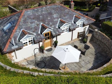 An outdoor patio with stone flooring a white umbrella two chairs a hanging chair and a building with dormer windows and a wooden door surrounded by stone walls at The Coach House at Lackington Farm in Piddletrenthide