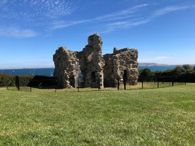Ruins of a stone structure surrounded by a black fence with grass in the foreground and sea in the background