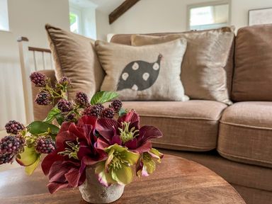 A close up of flowers on a wooden table with a sofa and a cushion with a bird design in the background at The Coach House at Lackington Farm in Piddletrenthide