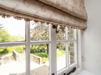A window with patterned fabric blinds looking out to a garden with trees and stone wall at The Coach House at Lackington Farm in Piddletrenthide