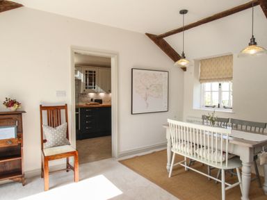 A dining area with a table and chairs next to a window and a wooden chair with a pillow near a doorway to a kitchen at The Coach House at Lackington Farm in Piddletrenthide