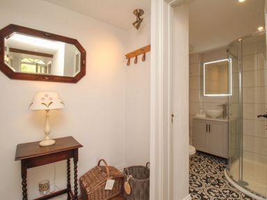 A small table with a lamp and mirror above it next to baskets and a doorway leading to a bathroom with a shower and patterned floor tiles at The Coach House at Lackington Farm in Piddletrenthide