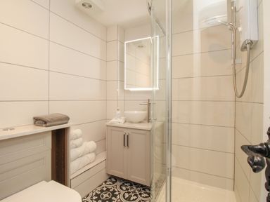 A bathroom with a shower, a vanity with a sink and mirror, folded towels on shelves, and patterned floor tiles at The Coach House at Lackington Farm in Piddletrenthide
