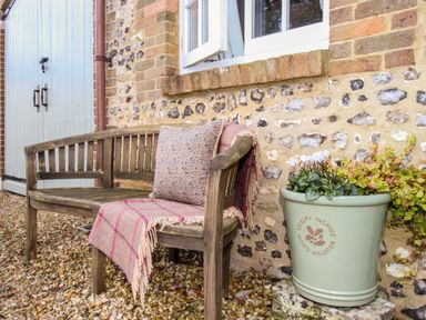 A wooden bench with a cushion and throw beside a stone wall with a plant pot outside The Coach House at Lackington Farm in Piddletrenthide