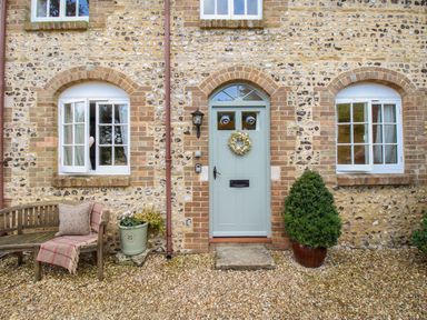 A stone house exterior with a sage green front door a wooden bench with a cushion and blanket and potted plants at The Coach House at Lackington Farm Piddletrenthide
