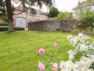 A garden with pink and white flowers green grass a tree stone walls and a brick building with gray doors at The Coach House at Lackington Farm in Piddletrenthide