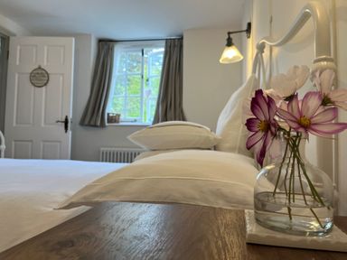 A bedroom with a bed and pillows a small vase with flowers on a wooden bedside table and a window with curtains at The Coach House at Lackington Farm in Piddletrenthide