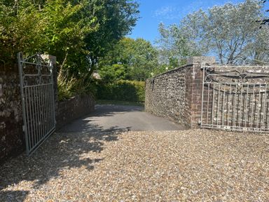 A gravel driveway with open wrought iron gates and stone walls surrounded by trees and bushes at The Coach House at Lackington Farm in Piddletrenthide