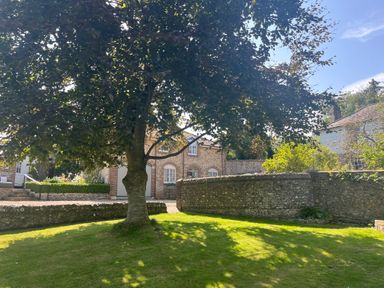 A garden with a large tree and a stone wall with a brick building in the background at The Coach House at Lackington Farm in Piddletrenthide