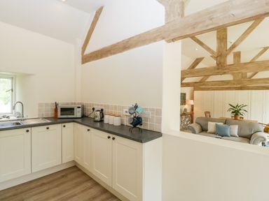 A kitchen with white cabinets a microwave kettle and mugs next to a living room with wooden beams a sofa and a plant at The Shippon in Honiton