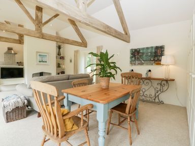 A dining area with a wooden table and chairs next to a living room with a gray sofa and exposed wooden beams at The Shippon in Honiton