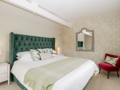 A bedroom with a green upholstered bed and a red chair with pillows a large mirror on the wall at The Shippon in Honiton