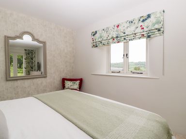 A bedroom with a bed covered by a light green blanket a window with floral curtains a red chair a wall mirror at The Shippon in Honiton