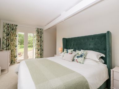 A bedroom with a large bed featuring a green headboard and floral cushions next to windows with floral curtains at The Shippon in Honiton