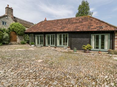A cobblestone courtyard with potted plants in front of a single story building with multiple glass doors and a tiled roof at The Shippon in Honiton