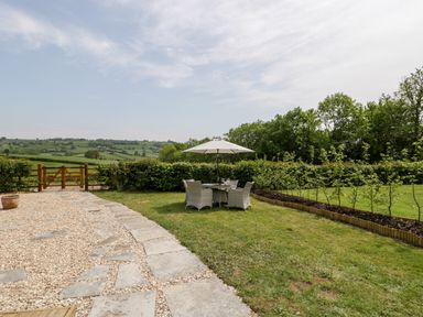 A garden area with a stone and gravel path a table with four chairs and an umbrella surrounded by hedges and trees at The Shippon in Honiton