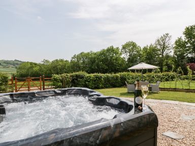 An outdoor hot tub with a bottle of champagne and two glasses beside it and a seating area with an umbrella in a garden at The Shippon in Honiton