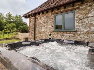 An outdoor hot tub next to a stone wall building with a window and a bottle with glasses on the edge at The Shippon in Honiton