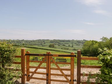 A wooden gate opening to a green field with benches and rolling hills in the distance at The Shippon in Honiton