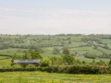 A rural landscape with green fields trees and a barn with solar panels on its roof at The Shippon in Honiton