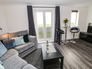 A living room with a grey sofa a black coffee table with a wine bottle and glass two black bar stools a round glass table with flowers and a kettle and a television at Sandy Corner in Blyth Northumberland