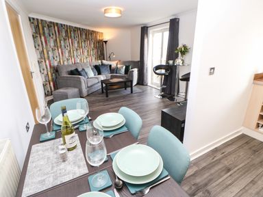 A dining table set with plates glasses and a bottle in a room with a grey corner sofa coffee table and bar stools at Sandy Corner in Blyth Northumberland