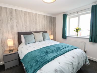 A bedroom with a bed featuring cushions and a blue throw a bedside table with a lamp and a window with curtains and flowers on the sill at Sandy Corner in Blyth Northumberland
