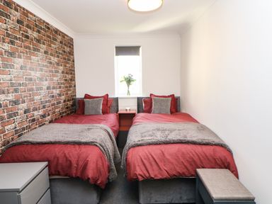 A bedroom with two single beds with red bedding and gray cushions a brick accent wall and a window with flowers on the windowsill at Sandy Corner in Blyth Northumberland