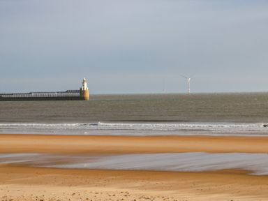 A sandy beach with waves and a lighthouse on a pier with a wind turbine in the sea at Sandy Corner in Blyth Northumberland