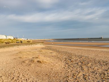 A sandy beach with a seawall benches and a pier in the distance at Sandy Corner in Blyth Northumberland
