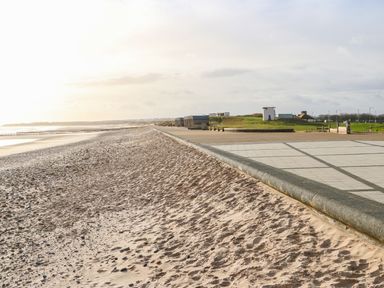 A sandy beach with a paved walkway and grassy area with buildings in the background at Sandy Corner in Blyth Northumberland