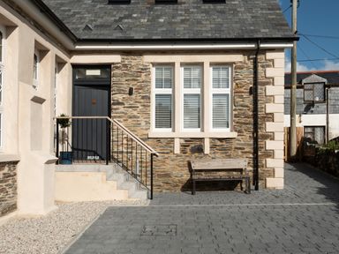 An exterior view of a stone house with three windows, a black door accessed by stairs, a bench, and a paved driveway at Church Rooms in Port Isaac