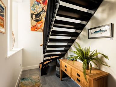 A hallway with a black metal staircase a wooden sideboard with a green plant and artwork on the walls at Church Rooms in Port Isaac
