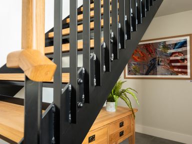 A wooden staircase with black metal railings next to a wooden table with a potted fern and a framed crab artwork on the wall at Church Rooms in Port Isaac