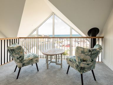 A loft area with two floral armchairs and a round wooden table by a large window overlooking rooftops and the sea at Church Rooms in Port Isaac
