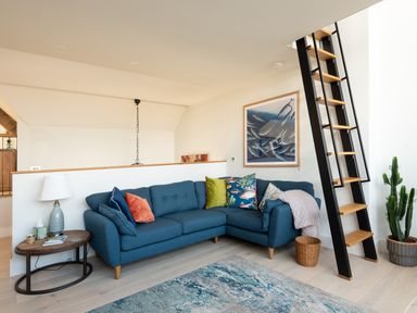 A living room with a blue sectional sofa cushions a ladder and a side table with a lamp at Church Rooms in Port Isaac