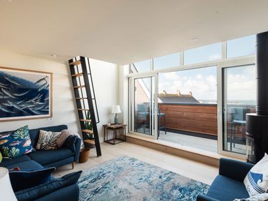 A living room with blue sofas a patterned rug a side table with a lamp a ladder leading to a loft and sliding glass doors opening to a balcony at Church Rooms in Port Isaac
