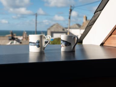 Two white mugs with sea life prints on a table outdoors with sea and buildings in the background at Church Rooms in Port Isaac