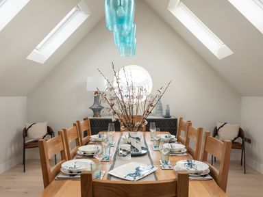 A dining room with a wooden table set with plates glasses and a vase of branches under blue hanging lights at Church Rooms in Port Isaac