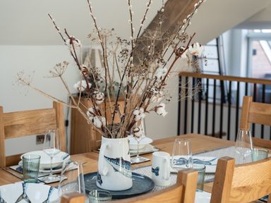 A dining table with wooden chairs set with plates glasses and a vase of dried flowers at Church Rooms in Port Isaac