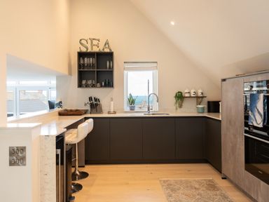 A kitchen with dark cabinets a window with a view of the sea and bar stools at Church Rooms Port Isaac
